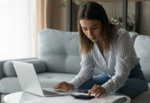 Studiengebühren im Check: Deine Rechte und Möglichkeiten Eine Studentin sitzt auf dem Sofa vor dem Laptop und berechnet am Taschenrechner Semesterbeitrag und Studiengebühren.