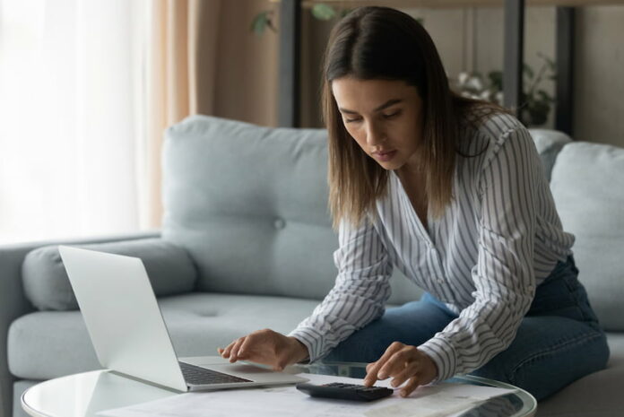 Eine Studentin sitzt auf dem Sofa vor dem Laptop und berechnet am Taschenrechner Semesterbeitrag und Studiengebühren.