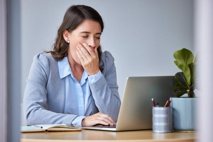 Eine Frau sitzt bei der Arbeit vor dem Laptop und gähnt. Sie leidet unter Frühjahrsmüdigkeit.