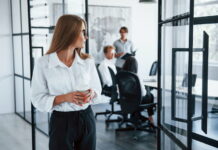 Mutig werden im Job und Alltag: Dein Weg aus der sozialen Phobie Eine Angestellte steht vor einem Büroraum, in dem sich Kollegen von ihr befinden, und schaut hinein. Sie hat eine soziale Phobie und ist distanziert.