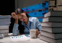 Wie viel Arbeit ist gesund? Zwischen Leistung und Lebensqualität Eine Frau sitzt müde mit einem Kaffee in der Hand am Schreibtisch, auf dem sich Ordner und Bücher stapeln, und stützt ihren Kopf mit einer Hand. "Wie viel Arbeit ist gesund?", fragt sie sich.