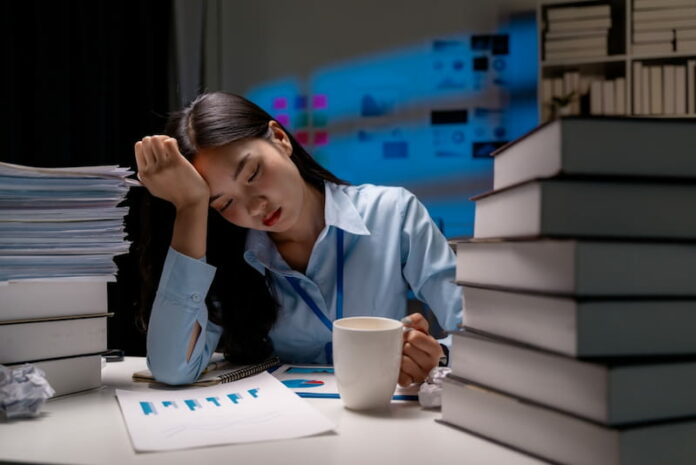 Eine Frau sitzt müde mit einem Kaffee in der Hand am Schreibtisch, auf dem sich Ordner und Bücher stapeln, und stützt ihren Kopf mit einer Hand. "Wie viel Arbeit ist gesund?", fragt sie sich.