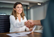 Unbefristeter Vertrag: Alles, was du wissen musst Eine Mitarbeiterin sitzt am Tisch und schüttelt lächelnd die Hand ihres Chefs. Ihr wurde ein unbefristeter Arbeitsvertrag vorgelegt.