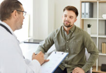 Gesundschreibung: Kann der Arbeitgeber sie verlangen? Ein Mann sitzt lächelnd vor seinem Arzt, der ein Klemmbrett in der Hand hält. Er ist für eine Gesundschreibung in der Praxis.