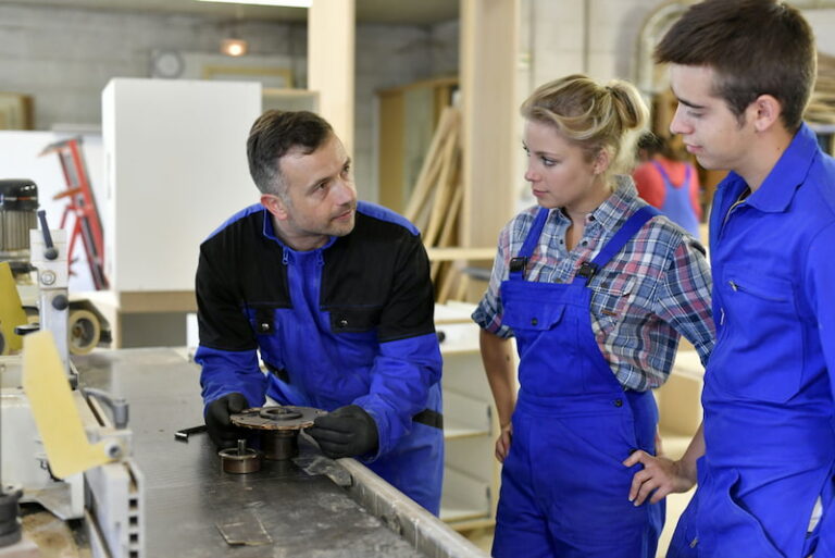 Zwei Auszubildende stehen vor einer Holzbearbeitungsmaschine und lassen sich von ihrem Berufsschullehrer die Funktion erklären. Als Azubis gilt für sie das Jugendarbeitsschutzgesetz.