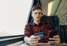 Kreditkarte für Studenten: So findest du die beste Lösung Ein junger Student sitzt im Zug. Er hält ein Smartphone und seine Kreditkarte für Studenten in den Händen.