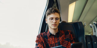 Kreditkarte für Studenten: So findest du die beste Lösung Ein junger Student sitzt im Zug. Er hält ein Smartphone und seine Kreditkarte für Studenten in den Händen.