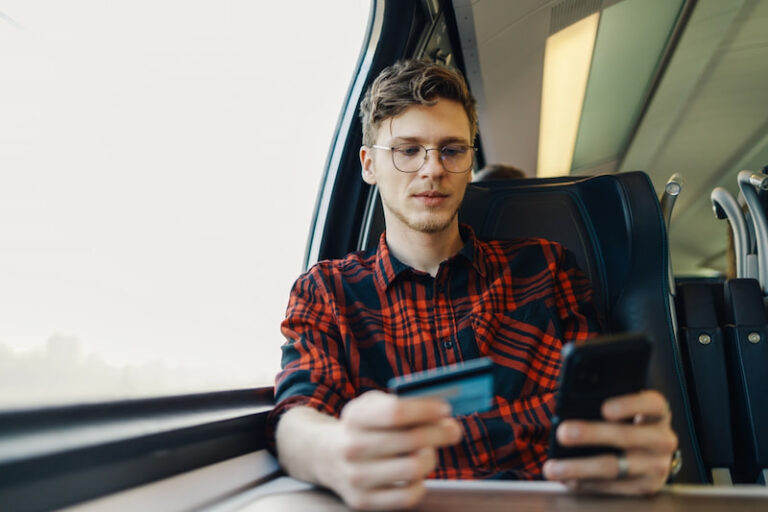 Ein junger Student sitzt im Zug. Er hält ein Smartphone und seine Kreditkarte für Studenten in den Händen.